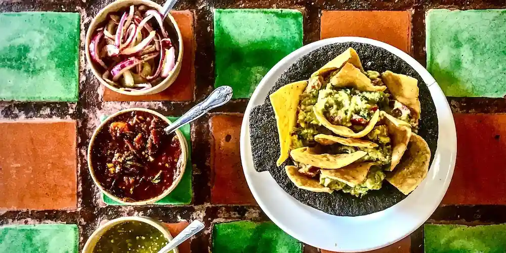 Tacos topped with guacamole beside bowls of salsa and pickled vegetables on a colorful tiled table in Mexico City